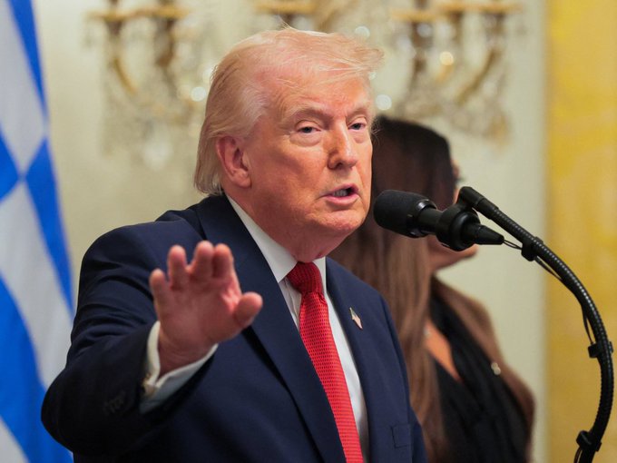 Donald Trump is seen speaking into a microphone at a formal event, gesturing with his hand, with a Greek flag visible in the background.