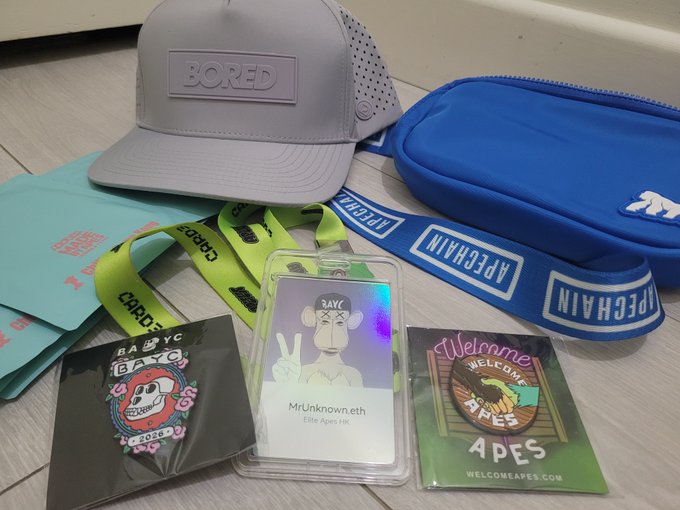 A Bored Ape Yacht Club-themed merchandise haul including a gray "BORED" cap, an ApeChain blue crossbody bag and lanyard, a "MrUnknown.eth" Elite Apes HK card, a 2020 BAYC pin, and a "Welcome Apes" sticker from WelcomeApes.com. The collection showcases the BAYC and Welcome Apes community swag received as a souvenir.