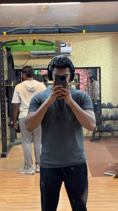 A mirror selfie of a man in a gym wearing over-ear headphones and a dark blue t-shirt, with gym equipment and another person visible in the background.
