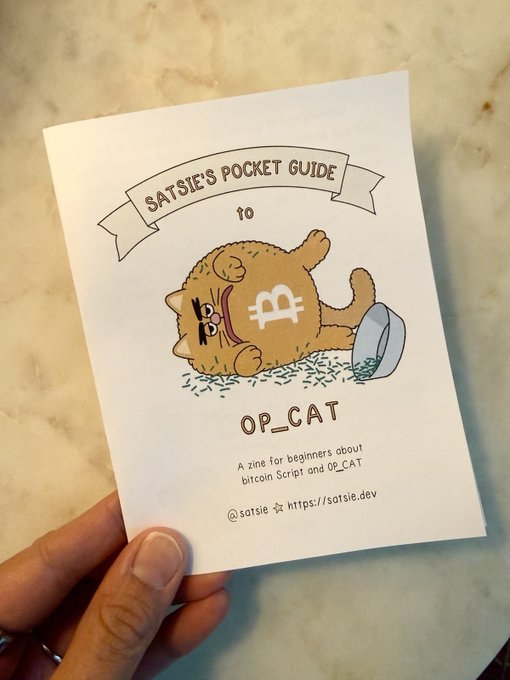 A hand holds a zine titled 'Satsie's Pocket Guide to OP_CAT' featuring an illustration of a cat with a Bitcoin symbol and a tipped-over fishbowl, described as 'A zine for beginners about bitcoin Script and OP_CAT.'