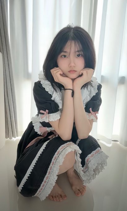 A young woman with short black hair wearing a black and white maid-style dress with pink bows, sitting barefoot on a white chair with her chin resting on her hands.