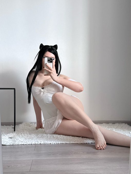 A cosplayer with black cat ears taking a mirror selfie, wearing a white strapless dress with lace details, seated on a white fluffy rug.