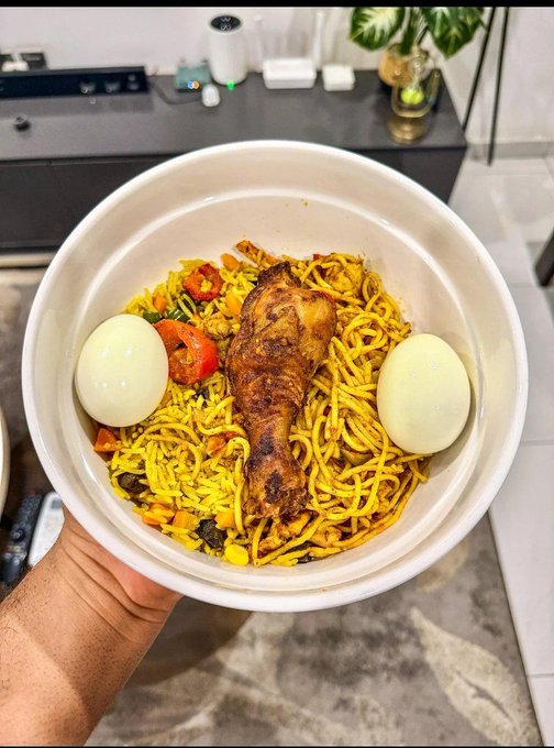 A hand holding a white bowl filled with yellow noodles, a chicken drumstick, two boiled eggs, tomatoes, and mixed vegetables.