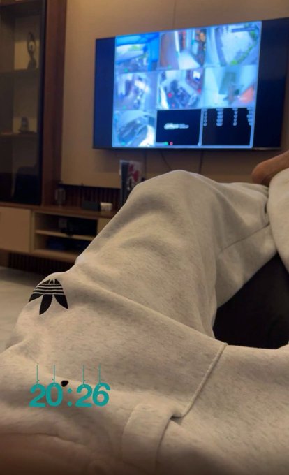 A cozy photo of someone lounging in a gray hoodie watching a surveillance monitor display on TV, with a timestamp of 20:26 in the corner.