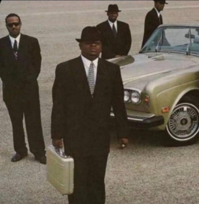 A man in a black suit, tie, and hat walks carrying a silver briefcase, followed by other men in suits, next to a luxury car.