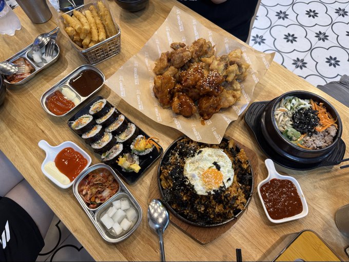 An overhead shot of a Korean meal spread including fried chicken, kimbap rolls, a fried rice dish with an egg, bibimbap, fries, and various dipping sauces on a wooden table.