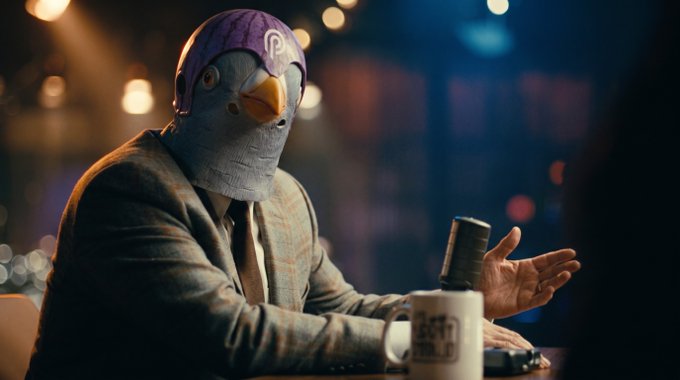 A man in a suit wearing a colorful pigeon mask sits at a table with a microphone and mug that reads 'THE LATE LATE SHOW WITH JAMES CORDEN'.