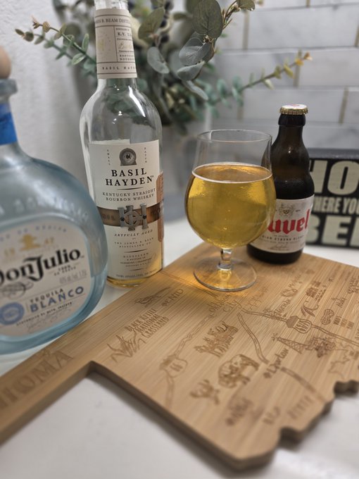 Three alcohol bottles—Don Julio Blanco tequila, Basil Hayden's bourbon, and Duvel beer—sit on a "Greatest Hits" wooden cutting board alongside a glass of amber beer, symbolizing a celebratory toast to three years of success with the @TENETTRADEGROUP.