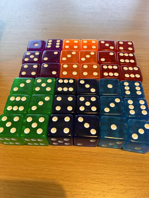 Twenty-four translucent dice in colors of purple, orange, red, green, black, and blue are arranged in a grid on a wooden table.