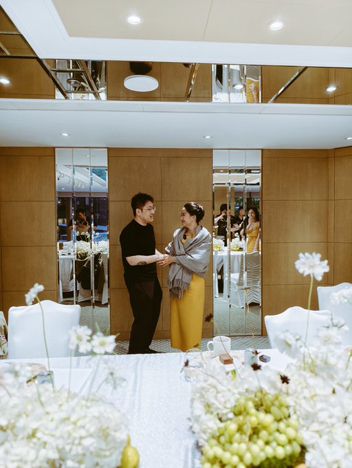 The image features two people standing next to each other, possibly at an event or gathering. They are positioned near a dining table with white flowers and grapes as decorations. The background of the photo shows several chairs arranged around the room, suggesting that this could be a social event or a party setting.