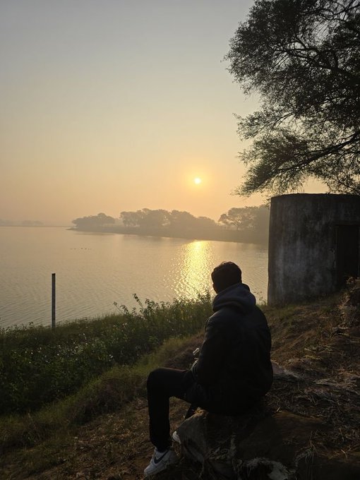 A serene photograph of a person in a dark jacket sitting by a body of water, watching the sun set over a misty horizon.