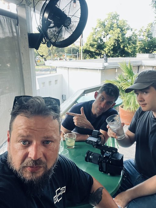 Selfie of three men sitting at an outdoor table under a fan, with one holding a drink and a camera placed on the table.