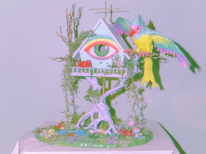 A whimsical, colorful sculpture features a house with a rainbow-colored eye on its facade, perched on a lavender tree with a vibrant parrot flying beside it, set on a grassy base with flowers.