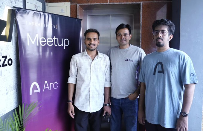 Three young men posing at an Arc Architects Meetup in front of a purple branded banner, with two of them wearing shirts featuring the Arc logo.