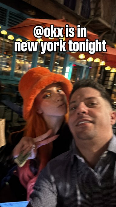 A selfie of a woman wearing an orange fluffy bucket hat and a man, with text overlay '@okx is in new york tonight', suggesting OKX cryptocurrency exchange promotional content.