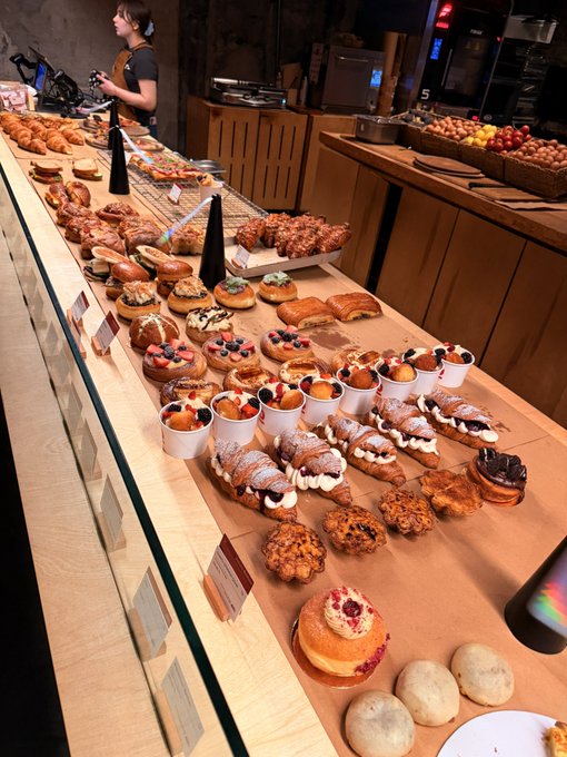 The image shows a long wooden counter displaying an assortment of pastries and donuts for sale at a bakery or cafe. There are various types of baked goods such as doughnuts, muffins, croissants, and cakes arranged on the counter in different sections. The display is well-organized and visually appealing, showcasing the wide variety of options available to customers. The image also includes text that reads "gm fam, just vibes", which could be a reference to the bakery's atmosphere or vibe.