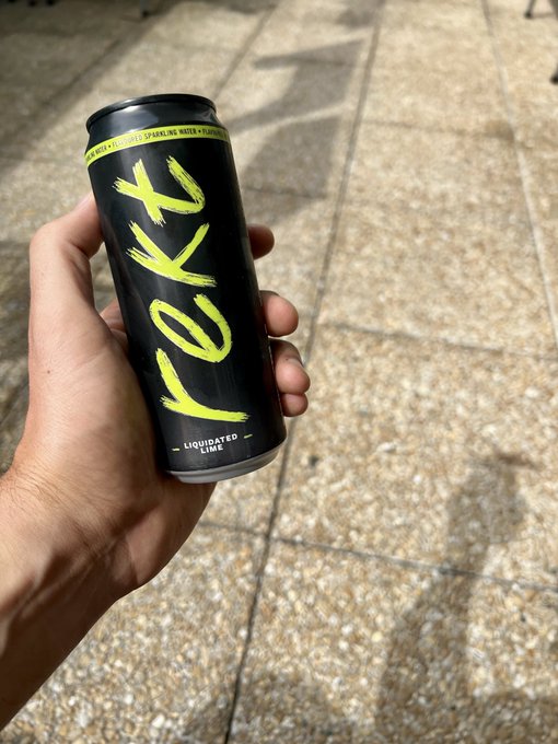 A hand holding a black can of 'LOXY' sparkling water, specifically the 'LIQUIDATED LIME' flavor, with green text on the side, standing over a tiled outdoor floor.