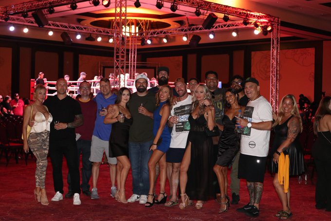 A diverse group of well-dressed attendees poses together for a group photo at what appears to be an upscale event venue featuring a boxing ring, dramatic stage lighting, and a red carpet. One person in the center wears a shirt reading "13.6K," suggesting this may be a milestone celebration. The caption "@synseerlii" and the greeting-style message indicate this is a social gathering where the host is sharing good vibes with their community or followers.