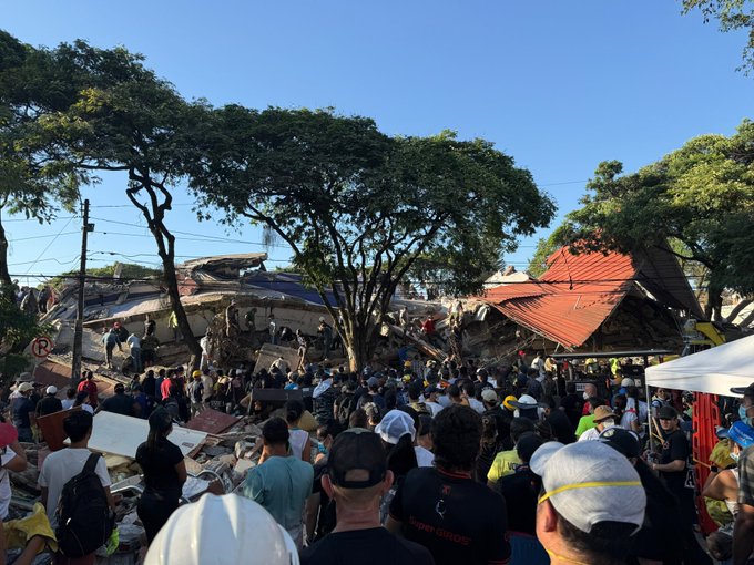 A large crowd gathers at the site of a collapsed building with red-tiled roofs under a clear blue sky, with rescue workers in helmets responding to the disaster.