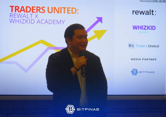 A man in a suit speaks into a microphone during a 'TRADERS UNITED: REWALT X WHIZKID ACADEMY' event, with the 'BITPINAS' and 'tru Traders United' logos displayed on the screen behind him.