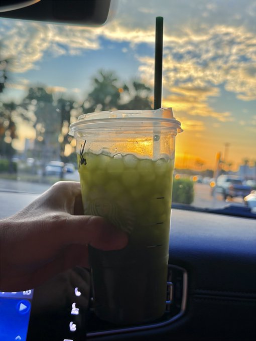 A hand holds an iced matcha green tea boba drink in a clear cup with a green straw against a backdrop of a cloudy sunset sky viewed from inside a car.