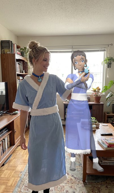 A real person cosplaying as Katara from Avatar: The Last Airbender in a blue and white outfit, standing next to a life-sized cardboard cutout of Katara holding her waterbending necklace.