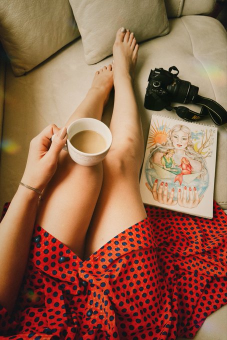 A first-person view of someone relaxing on a couch, holding a coffee mug, with their bare feet visible next to a Canon camera and an illustrated notebook, wearing a red polka dot skirt.