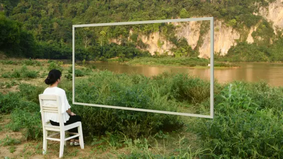 A person in a white shirt sits on a white chair facing a large white rectangular frame that overlooks a river and lush green mountains, creating a window-like view of the landscape.