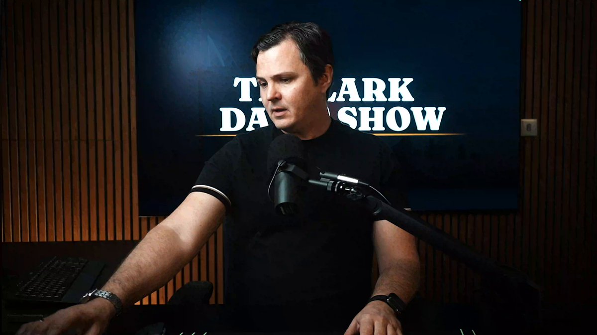 A man wearing a black shirt is sitting at his computer, with the words "The Clark Show" written above him. He has a microphone placed next to him as if he's preparing for or currently recording a podcast. Above him on the wall, there are several charts displayed which might be related to Bitcoin and cryptocurrency. The image also includes text that mentions Bitcoin shorts, Bitcoin hopium, Bitcoin coolers, and software stocks in a discussion about Bitcoin and the stock market. Additionally, a social media account logo is visible at the bottom of the photo.