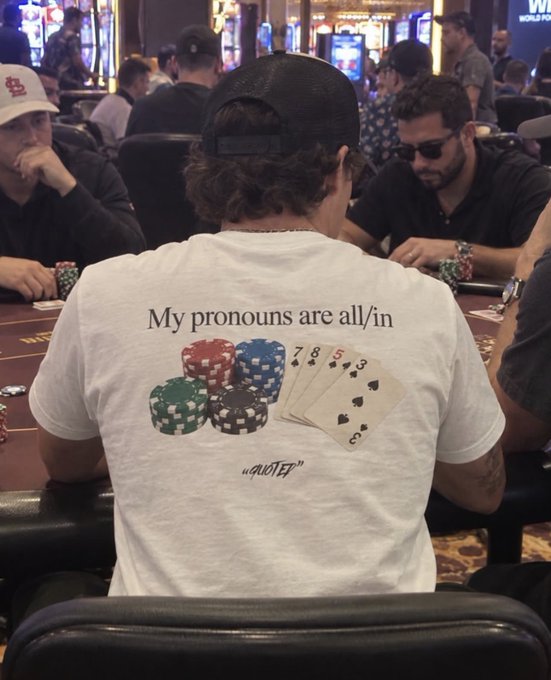 A poker player sits at a table wearing a white t-shirt that reads "My pronouns are all/in" with poker chips and playing cards, ending with "QUOTE" at the bottom.