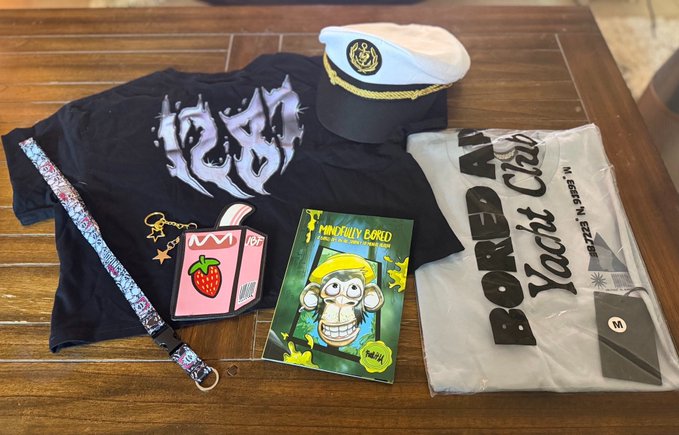 A flat lay on a wooden table showing a navy t-shirt with a tribal-style graphic, a white captain's hat with gold detailing, a patterned lanyard, a pink juice box-style card, a 'Mintyly Bored' monkey NFT card, a 'BORED APE Yacht Club' plastic bag, and a black phone case.