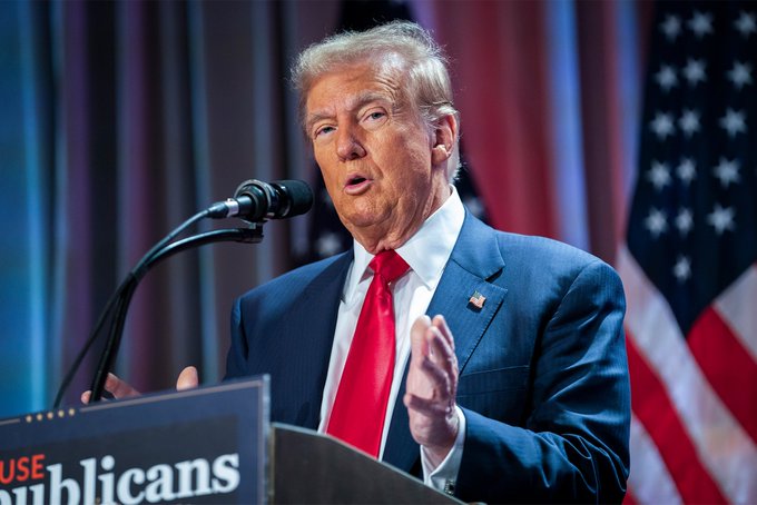 Donald Trump, wearing a blue suit and red tie, speaks at a podium with a microphone. The podium displays partial text reading 'USE' and 'ublicans', with an American flag visible in the background.