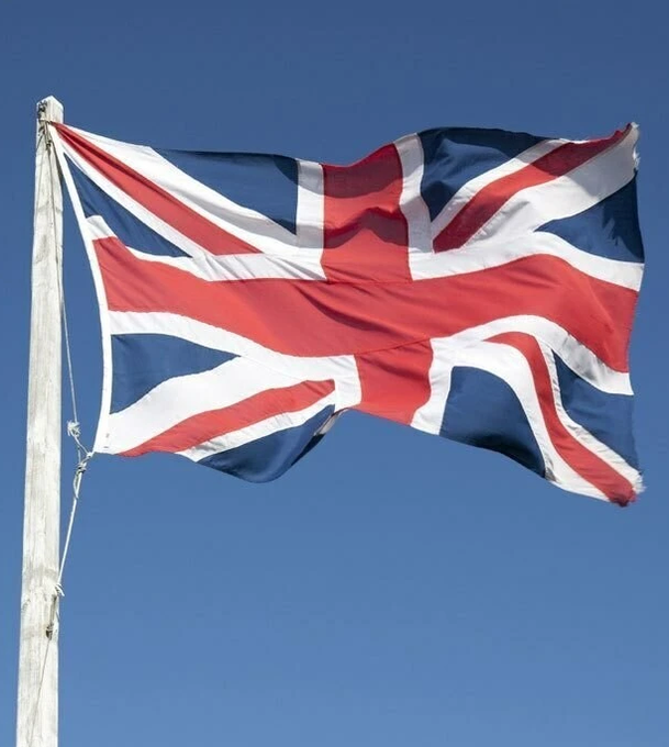 A weathered British Union Jack flag waves against a clear blue sky, mounted on a wooden flagpole. The image accompanies news about U.K. crypto tax disclosures, symbolizing the British government's oversight of cryptocurrency gains among high-earning investors.