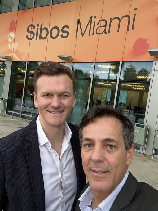 A selfie of two men in business attire standing in front of an orange building facade with the text 'Sibos Miami'.