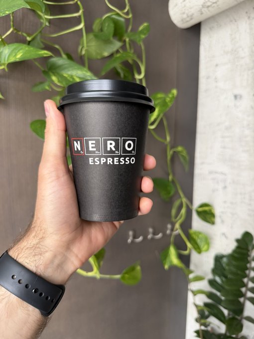 A hand holds up a black matte takeaway cup labeled "NERO ESPRESSO" against a backdrop of trailing green pothos vines, with a fitness tracker visible on the wrist. The post's reference to "Friday coffee" pairs perfectly with the caffeinated fuel in hand—just what one needs to power through the end of the workweek and "pretend we were productive all week."