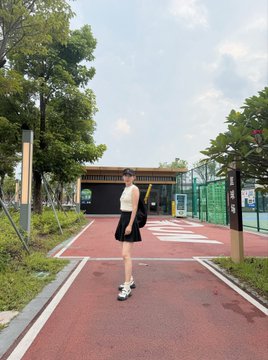 A young woman in athletic wear stands on a red running track holding what appears to be sports gear, with a large facility and green courts in the background. The image ties to the post by showcasing the stylish Binance-branded athletic apparel that inspired her to get active and head to the court despite the summer heat.