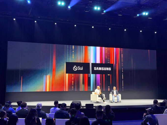 Two men sit on stage in white chairs at a conference, with a large screen behind them displaying the Sui and Samsung logos against a colorful, streaked background.