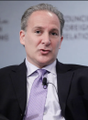 A man in a dark suit and purple tie speaks at what appears to be an investment conference, with partial text visible in the background suggesting a financial or economic discussion, fitting the post's theme comparing Bitcoin and gold as appreciating assets.
