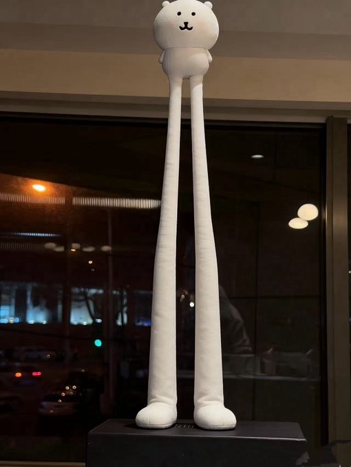 A tall white bear statue with elongated legs stands on a pedestal inside a building, displayed against a backdrop of windows reflecting nighttime city lights. The whimsical, towering figure complements the playful tone about thinking of loved ones and craving KFC on Thursdays.