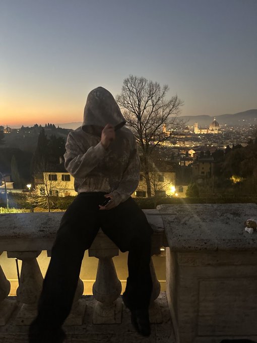 A hooded figure sits on a stone balustrade at dusk, silhouetted against a panoramic view of a city skyline with a prominent dome, likely Florence.