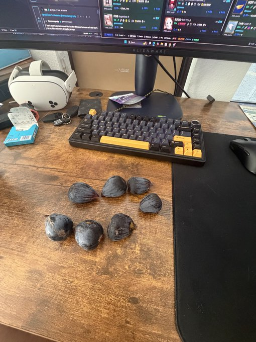 A desk setup featuring a monitor, keyboard, VR headset, and seven dark, round objects arranged on the wooden surface.
