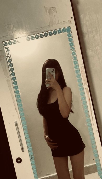A young woman with long dark hair, wearing a short black dress, takes a mirror selfie in what appears to be a bathroom with teal-tiled mirror framing, while holding her phone in front of her face. The image ties to the caption's lament about attractiveness not being enough to prevent getting dumped.
