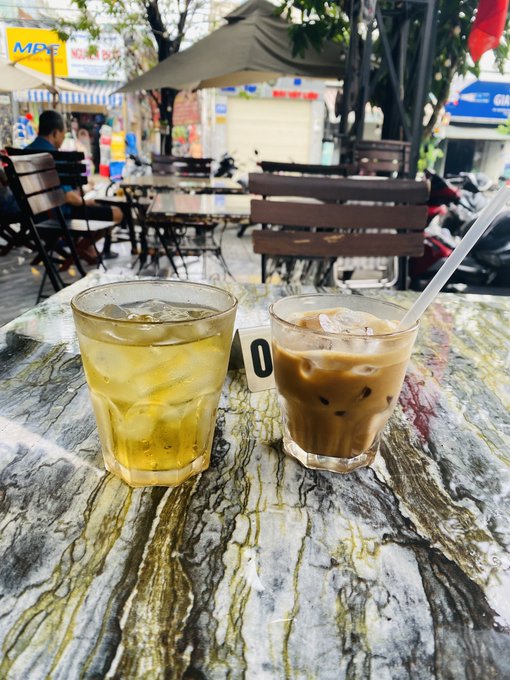 Two glasses of iced beverages—one green tea and one coffee with milk—sitting on a marble-patterned table at an outdoor Vietnamese café with an MPC sign visible in the background.