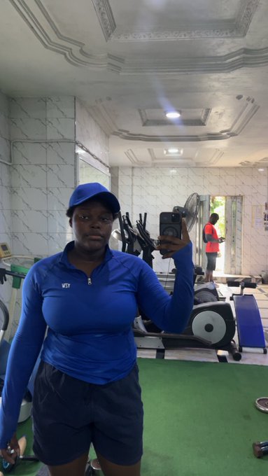 A woman in a blue long-sleeved athletic top, cap, and dark shorts takes a mirror selfie in what appears to be a gym or fitness center.