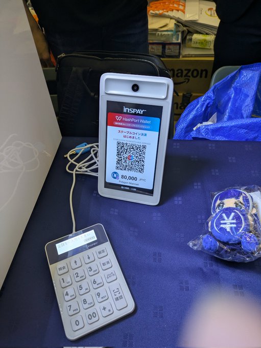 A JPYC payment terminal displays a QR code for 80,000 JPY on its screen, alongside a keypad and a small bag of ¥-symbol coins on a blue surface, illustrating the new crypto payment setup in action.