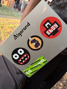 The image shows a person holding a silver laptop computer with stickers of three different cartoon characters, including one that looks like a woman wearing a dress. The laptop has the words "Algofam" written on it. The person appears to be in an outdoor setting, possibly at a gathering or event where people are standing and socializing around them.