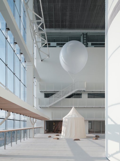 A modern architectural interior featuring a large white balloon-like sculpture suspended from the ceiling above a small white tent installation in a bright, minimalist atrium with floor-to-ceiling windows.