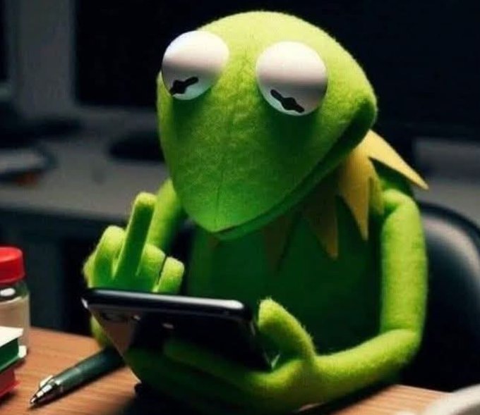 A Kermit the Frog plush toy sitting at a desk looking at a smartphone with half-closed eyes, positioned next to a pen.