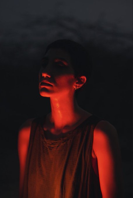 A dramatic portrait of a woman with a shaved head wearing a sleeveless top, illuminated by a strong red light from below against a dark, cloudy background.