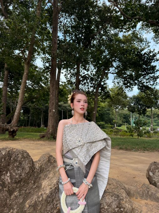 A woman wearing traditional Thai-style attire with a sash and ornate jewelry, standing outdoors in a park-like setting with large trees and rocks, posing gracefully.